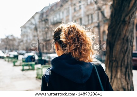 Young woman in black coat and with curly hair walking through an old European city street next to road and cars on autumn day, view from behind. Royalty-Free Stock Photo #1489920641
