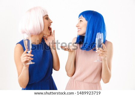Portrait of two excited women wearing blue and pink wigs talking while drinking champagne isolated over white background