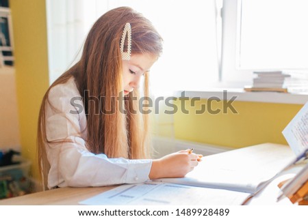 Education at home concept - Cute little girl with long hair studying or completing home work on a table with pile of books and workbook.