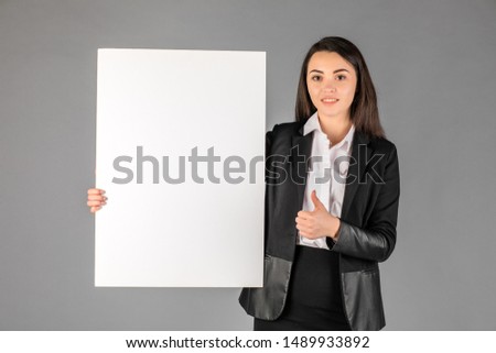 Young smiling woman pointing at a blank white sheet for advertisement on a gray background