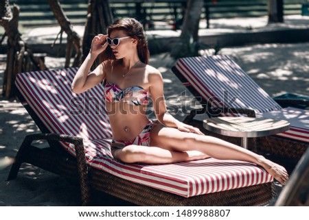 Beautiful young lady in glamorous sunglasses and cute bikini rests on the lounge on the sunny beach; recreation concept.