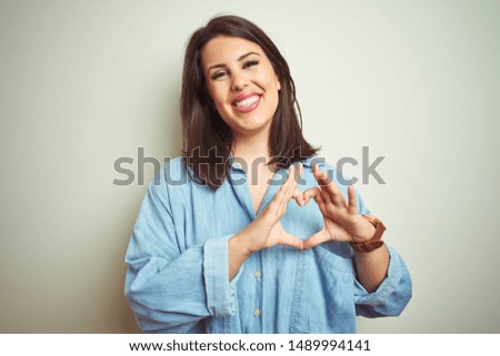 Young beautiful brunette woman wearing casual blue denim shirt over isolated background smiling in love showing heart symbol and shape with hands. Romantic concept.