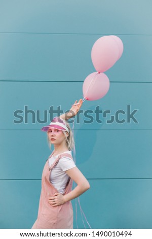 Blonde teen girl in cute pink clothes stands with balls in hands on blue background, looks in camera. Isolated. Copyspace. Vertical photo.
