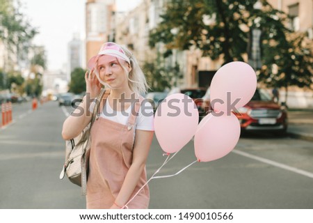 Cute girl in pink clothes and blond hair poses on the street with balloons in her hands, looks away and smiles on the background of cityscape. Portrait of a cheerful teen girl with balloons