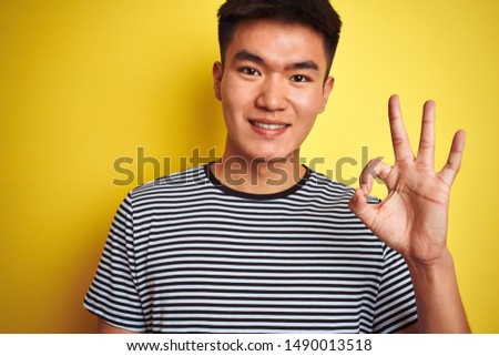 Young asian chinese man wearing striped t-shirt standing over isolated yellow background doing ok sign with fingers, excellent symbol