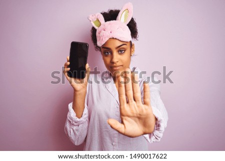 Afro woman wearing pajama and mask showing smartphone over isolated pink background with open hand doing stop sign with serious and confident expression, defense gesture