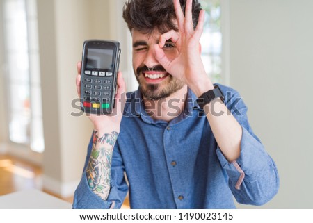 Young man holding dataphone point of sale as payment with happy face smiling doing ok sign with hand on eye looking through fingers