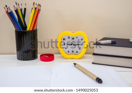 Stationery items. Colored pencils, books, pen, notebook and small alarm clock on the table.