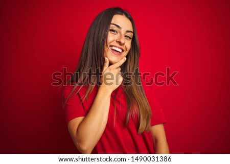 Young beautiful woman wearing t-shirt standing over isolated red background looking confident at the camera smiling with crossed arms and hand raised on chin. Thinking positive.
