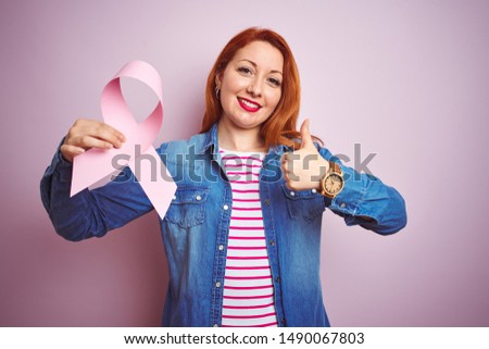 Young beautiful redhead woman holding cancer ribbon over pink isolated background happy with big smile doing ok sign, thumb up with fingers, excellent sign