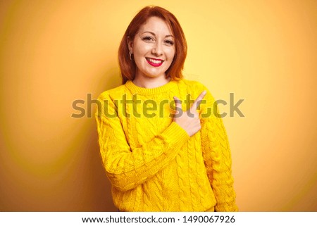 Beautiful redhead woman wearing winter sweater standing over isolated yellow background cheerful with a smile on face pointing with hand and finger up to the side with happy and natural expression