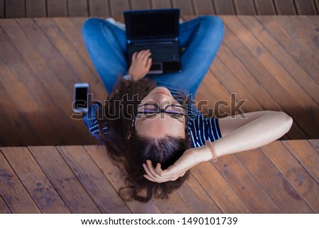 A tired brunette girl in glasses is sitting cross-legged on a bench, a laptop is on her lap, her face is raised up, one hand on her head. View strictly from above, outdoor.