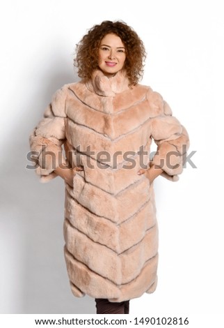 Smiling woman with long curly red hair poses demonstrating beige midi fur coat trimmed with her hands in her pockets on white background