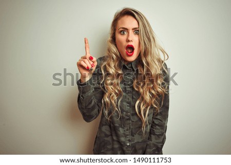 Young beautiful woman wearing military camouflage shirt over white isolated background pointing finger up with successful idea. Exited and happy. Number one.