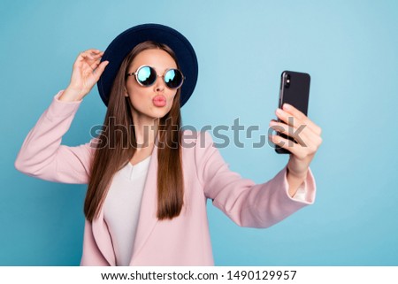 Portrait of dreamy cute girl using her mobile phone making selfie sending air kisses launching blog stream wearing pink stylish topcoat isolated over blue background