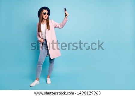 Full size photo of childish girl using her cell phone taking self photo having fun wearing eyeglasses eyewear topcoat denim jeans isolated over blue background
