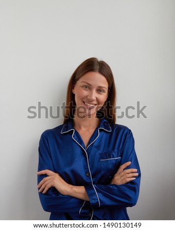 Brown parted hair woman with happy face and smile with green eyes in blue silk pajamas jacket holds hands cross together and looks to camera in morning in natural light on the white wall background  Royalty-Free Stock Photo #1490130149