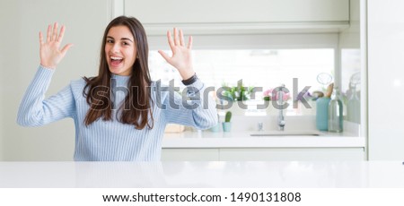 Wide angle picture of beautiful young woman sitting on white table at home showing and pointing up with fingers number ten while smiling confident and happy.