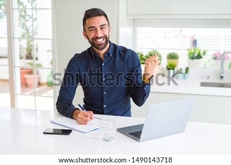 Handsome hispanic man working using computer and writing on a paper screaming proud and celebrating victory and success very excited, cheering emotion