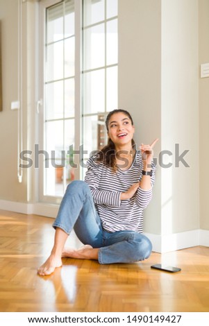 Young beautiful woman sitting on the floor at home with a big smile on face, pointing with hand and finger to the side looking at the camera.