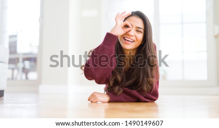 Young beautiful woman lying on the floor at home with happy face smiling doing ok sign with hand on eye looking through fingers