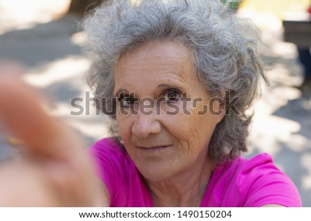 Positive old lady taking selfie in park. Senior grey haired woman in casual holding gadget with hand and looking at phone camera. Self shot concept