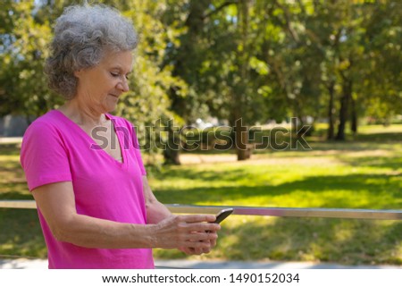 Positive focused old lady texting message while walking on parkway. Happy senior grey haired woman in casual using mobile phone in park. Wireless connection concept
