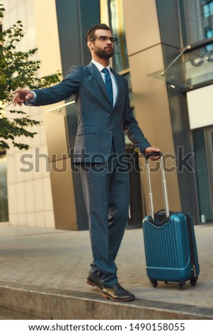 Catching taxi. Full length of young and handsome bearded man in suit carrying suitcase and raising his arm while standing outdoors