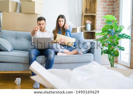 Young beautiful couple relaxing sitting on the sofa of new house using computer laptop pointing with finger to the camera and to you, hand sign, positive and confident gesture from the front