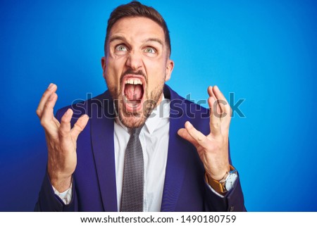 Close up picture of young handsome business man over blue isolated background crazy and mad shouting and yelling with aggressive expression and arms raised. Frustration concept.