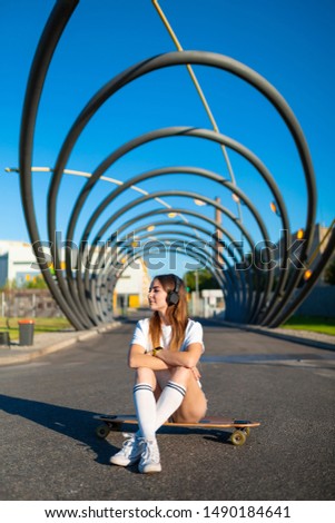 A girl in baseball cap sits on skateboard, longboard. In summer in city on background of an asphalt road, a young woman. In his hand a smartphone, an application to the Internet. Free space for text.