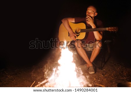 Hipster man smoking cigarette and holding acoustic guitar at big bonfire at camp, chilling at camp in the evening . Musician relaxing at fire , camping in woods. Copy space