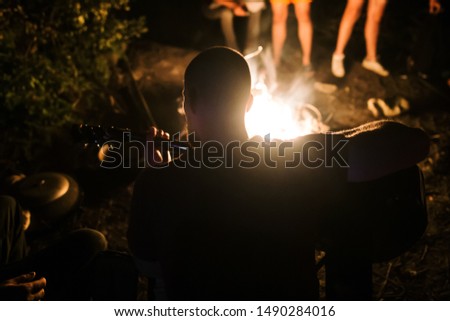 Hipster man playing on acoustic guitar and singing song at big bonfire at camp, chilling with friends in the forest. Group of people relaxing at fire in the evening, camping near lake