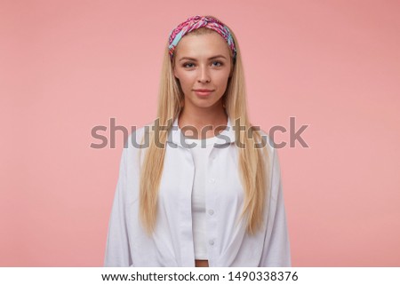 Beautiful young woman with long blond hair looking at camera with soft smile, wearing colored headband and white shirt, standing over the pink background
