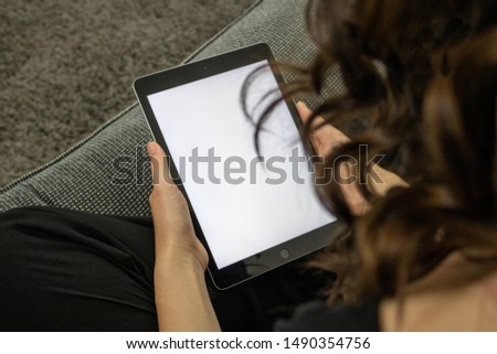 Young brunette woman playing on a tablet sat on a sofa relaxing, the tablet has a white full size screen to enable adding text or images. 