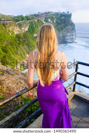 Blonde girl with long hair in traditional purple skirt at sunset on ocean background and Uluwatu temple, Bali, Indonesia
