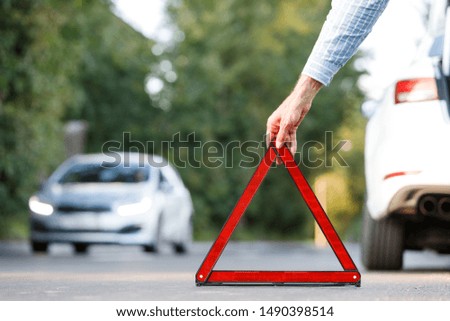 Close up of man driver putting red warning triangle/emergency stop sign behind his broken car on the side of the road, copy space and blurred car on background.