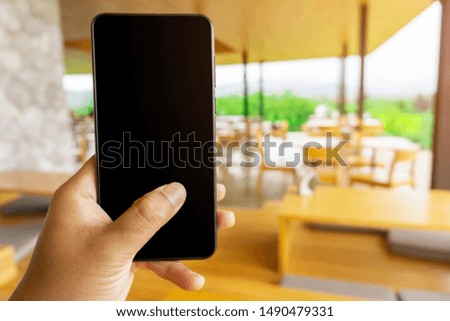 Close up young asian woman hand holding a modern black smartphone mock up in vertical position with blank screen against beautiful blurred coffee shop interior background.