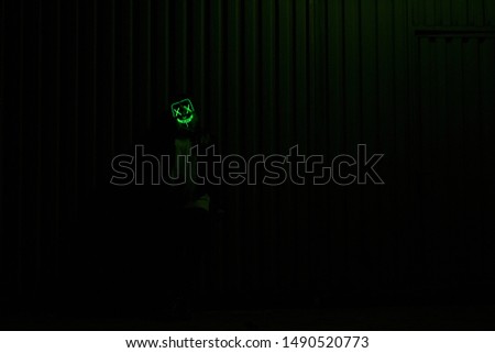 Model wearing scary green led halloween mask leaning against the metal wall in the dark
