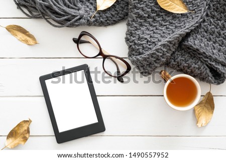 Autumn composition. Feminine desk table with knitted scarf, tea cup, glasses, tablet ebook with blank screen, fall leaves on wooden background. Flat lay, top view. Nordic, hygge, cozy home concept