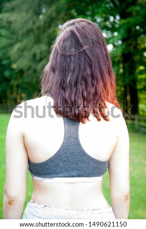 white girl's back and bare shoulders in grey vest style crop top with shoulder length reddish brown hair in a green outdoor setting