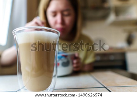 Woman on the kitchen is eating her breakfast and going to drink coffee. She dressed in yellow T-shirt and holding spoon in right hand. She is chewing and  in haste to her job. Local focus on the glass