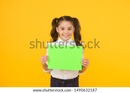 Here is your homework. Adorable small child holding empty homework sheet on yellow background. Cute little girl with blank green school paper for doing homework assignment. Homework, copy space.