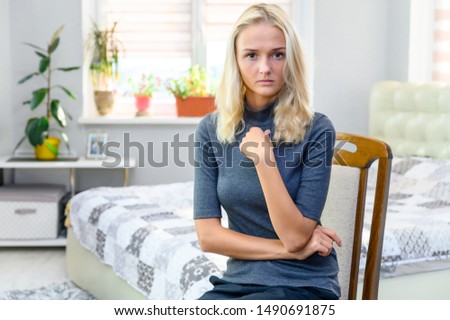Studio portrait of a pretty blonde student girl, a young woman in a gray sweater sitting on a chair in the room. Dissatisfied, bored.