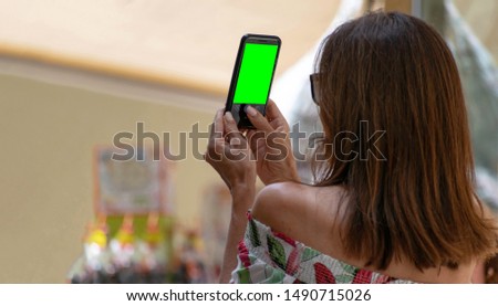 Close up rear view of a brown haired middle aged caucasian woman, wearing a colorful shirt, taking a picture with her smartphone on a sunny day with a blurry background.