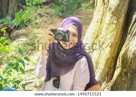 Portrait selective focus of young female tourist sitting by a tree in a forest while holding and taking photos with her retro film camera. 