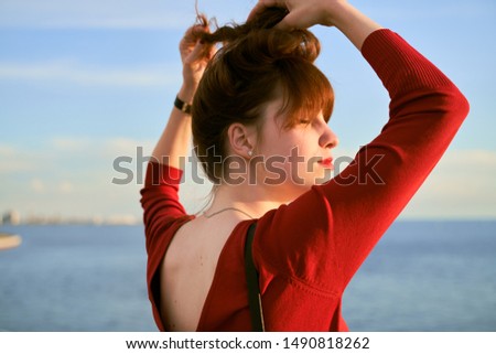 Ginger haired woman ties her long hair into knot. Photo of the girl back. The plunging neckline on red dress. The lady enjoys evening sea breeze standing on bridge.
