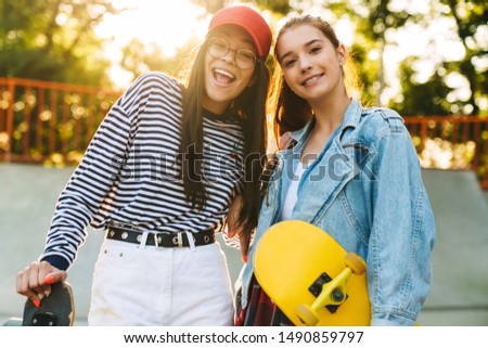 Image of two cute girls wearing casual clothes smiling and poising with skateboards in skate park