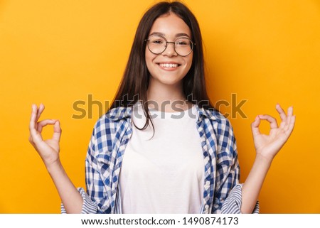Picture of a smiling young cute teenage girl in glasses meditate isolated over yellow wall background.