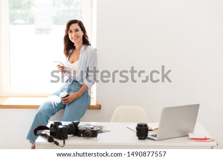Beautiful young woman photographer working at her office while sitting at the desk, using mobile phone